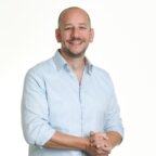 Een glimlachende man met een kaalgeschoren hoofd, gekleed in een lichtblauw overhemd met knopen.