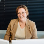 Een glimlachende vrouw in een blazer, met een headset op, zit achter haar computer in Huisartsenpraktijk Snijders.