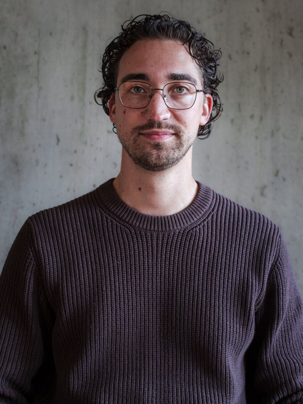 Een man met een bril en een donkere trui die naar de camera kijkt.