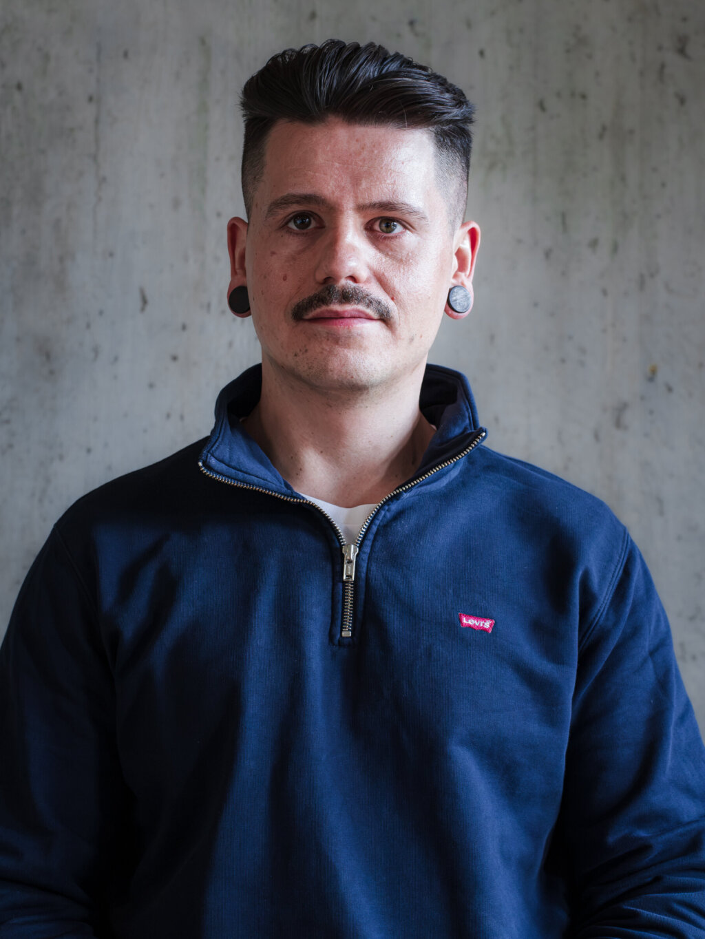 Een man met een snor en oorpiercings, gekleed in een blauw sweatshirt met kwartrits.