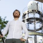 Een glimlachende man in een wit overhemd die omhoog kijkt naar een grote klokkentoren.